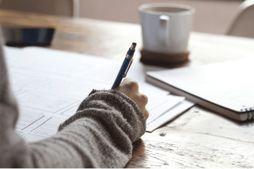 A person focused at their desk, pen in hand, surrounded by scattered papers, with a steaming cup of coffee placed nearby.