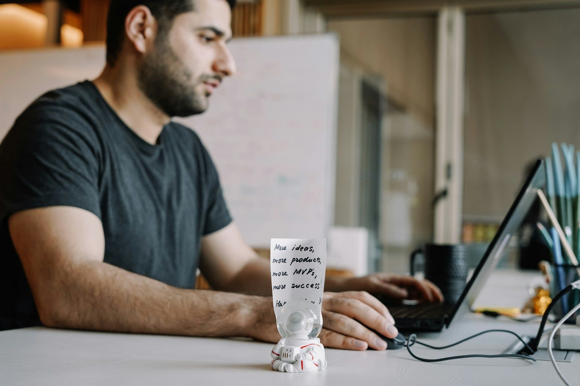 A business owner works at a laptop in an office with a small note of ideas next to him