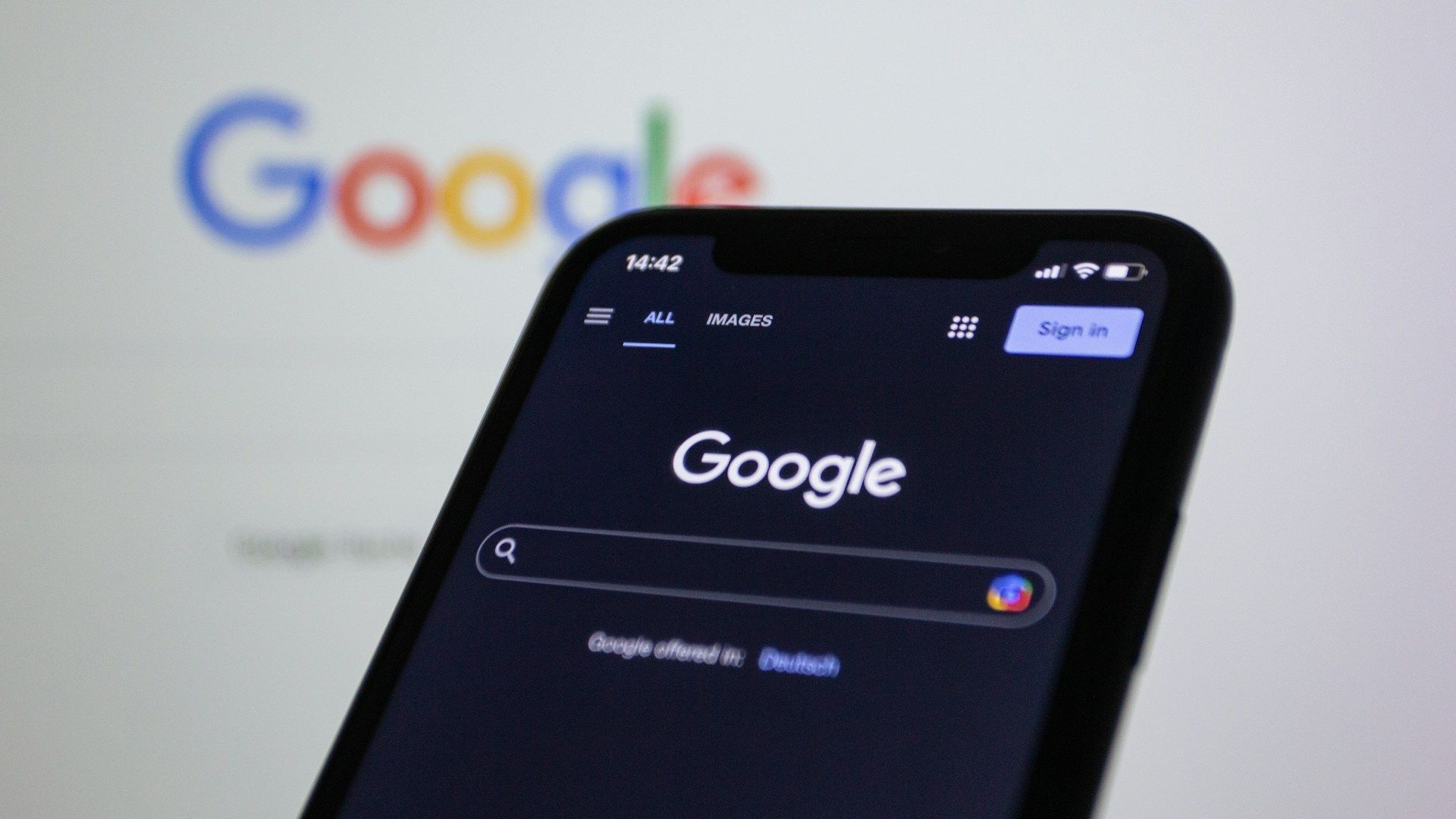 A smartphone with the Google homepage in dark mode in front of a computer screen